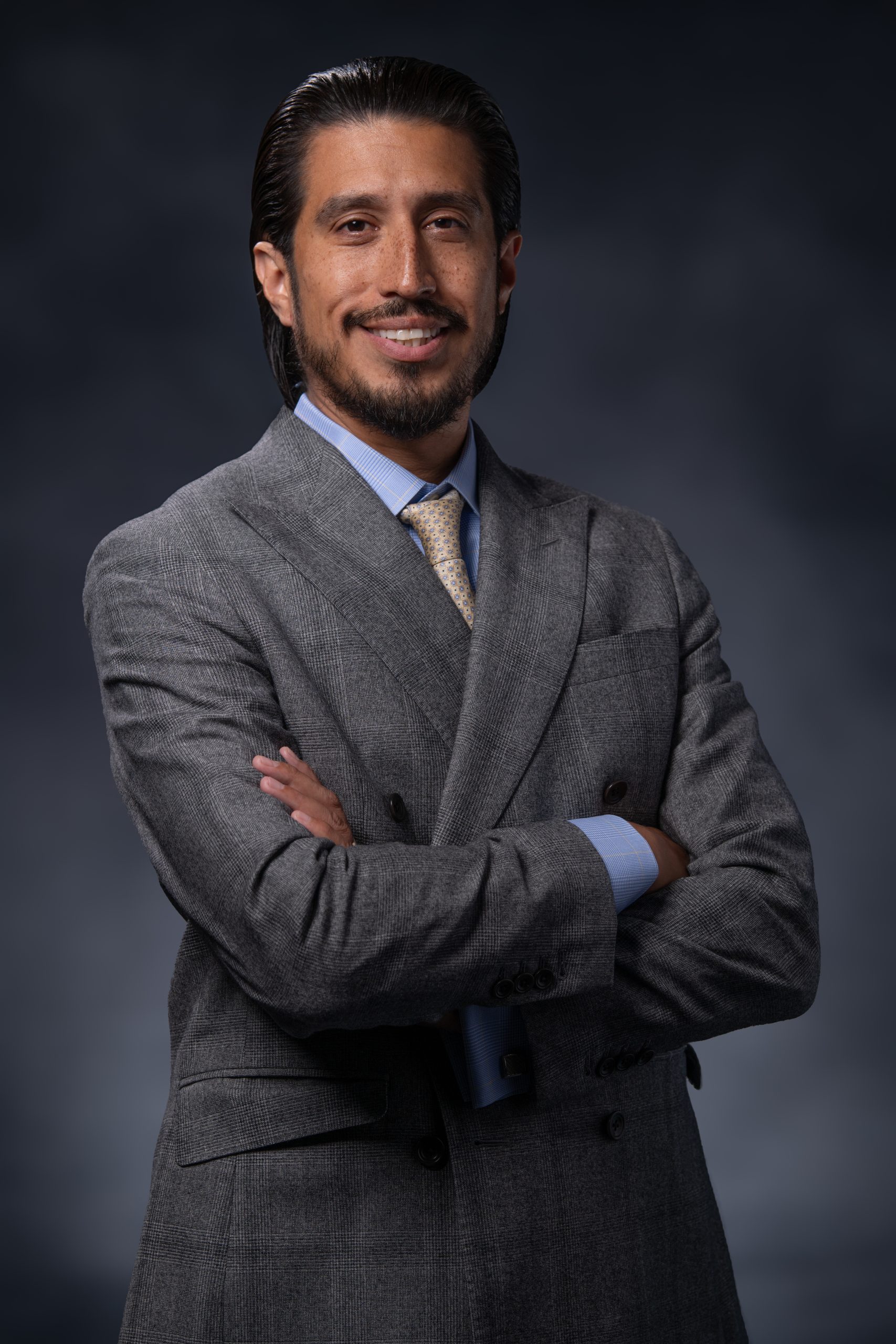 Professional portrait of a man with dark hair and a beard, in a gray suit and blue shirt, smiling at the camera.